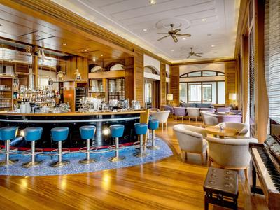 Bar d'hôtel confortable avec sol en bois, tabourets bleus et coin salon avec fauteuils près de grandes fenêtres.