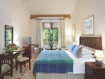 Chambre lumineuse avec grand lit, table, chaise et vue sur jardin verdoyant.