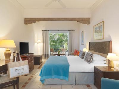 Helles Schlafzimmer mit Doppelbett, blauer Tagesdecke, Holzmöbeln und Terrasse mit grüner Aussicht.