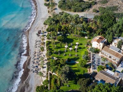 Grüner Park mit Palmen, Sonnenschirmen und Gebäuden neben klarer Küstenlinie mit blauem Meer.