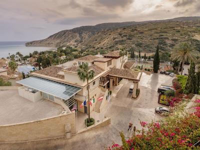Landschaft mit Hügeln, Palmen, Blumen und einem großen, beigefarbenen Gebäude nahe der Küste.