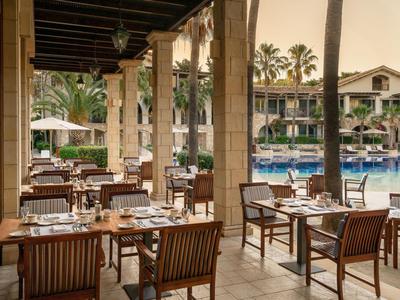 Espace couvert d'un restaurant extérieur avec des tables en bois donnant sur une piscine et des palmiers.