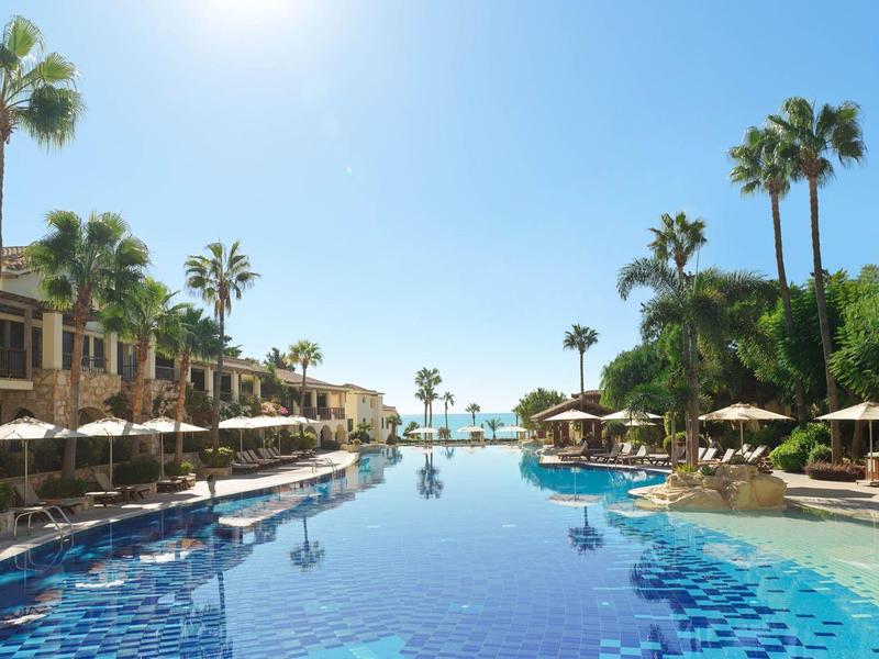 Großer blauer Pool mit Sonnenschirmen, Palmen und hellen Gebäuden unter klarem Himmel.