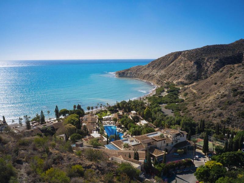 Küstenlandschaft mit blauem Meer, steilen Hügeln und einem kleinen Dorf mit Häusern und Bäumen.
