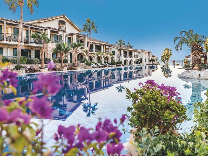 Großer blauer Pool vor einem Hotel mit Blumen und Palmen unter klarem Himmel.