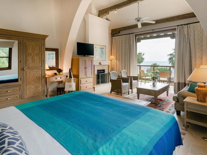 Hell eingerichtetes Schlafzimmer mit großem Bett, Holzmöbeln und Meerblick durch Fenster und Balkon.