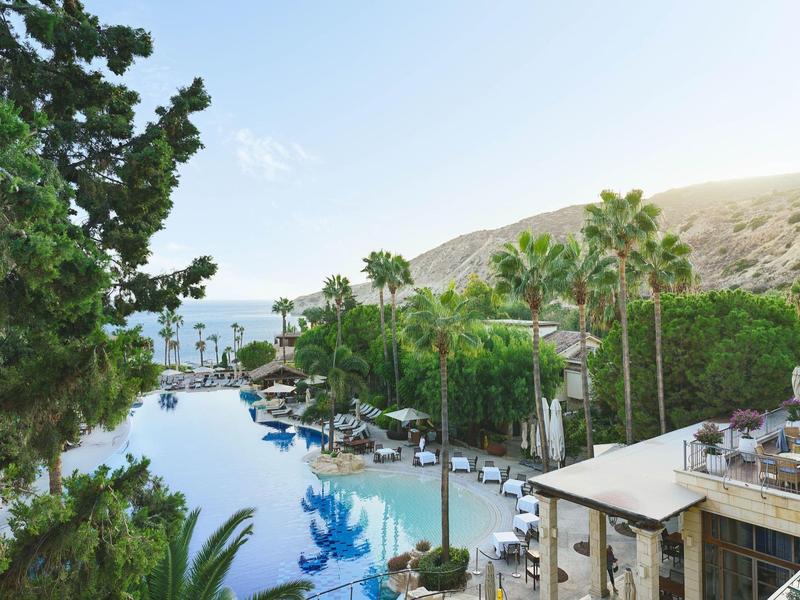 Großer Pool umgeben von Palmen, grünen Bäumen und Hügeln unter blauem Himmel.