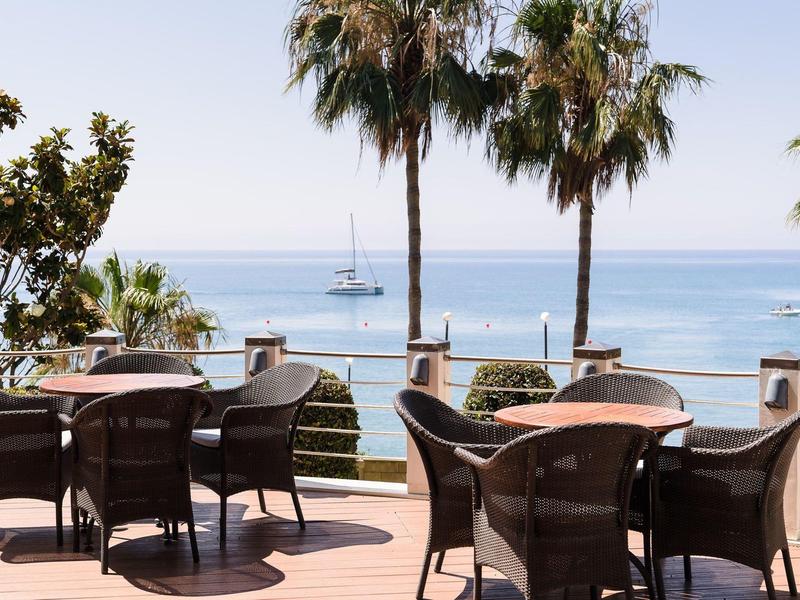 Leere Terrasse mit Tischen und Stühlen, Palmen und Meer im Hintergrund unter klarem Himmel.