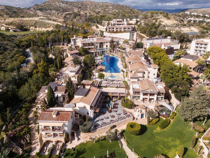 Luftaufnahme eines Resorts mit Pool, umgeben von Gebäuden, Bäumen und Hügeln im Hintergrund.