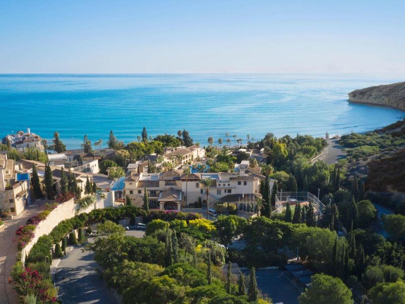 Küstenlandschaft mit Meer, Häusern, grünen Bäumen und Hügeln unter klarem blauem Himmel.