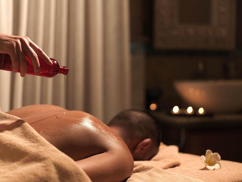 Person liegt auf Massagetisch, Hand gießt Öl auf Rücken, warmes Licht, ruhige Atmosphäre.