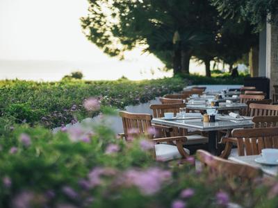Terrasse eines Hotels mit Holztischen und -stühlen neben grüner Vegetation und Blick aufs Meer.
