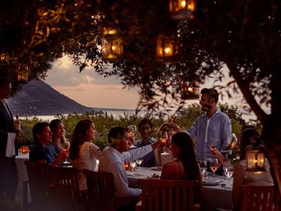 Abendessen im Freien am Meer mit Menschen, Laternen und Bergen im Hintergrund.