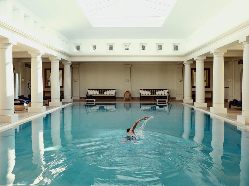 Großes Hallenbad mit Säulen, hellem Dachfenster und schwimmender Person in der Mitte des Beckens.