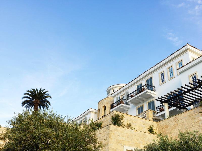 Großes weißes Gebäude mit Balkonen, blauer Himmel, Palmen und steinerne Mauer im Vordergrund.