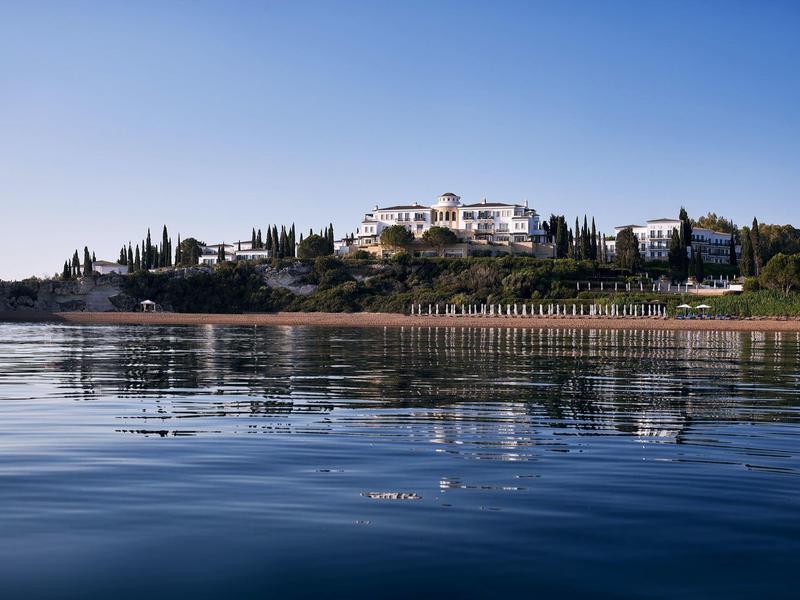 Weißes Gebäude auf grünem Hügel mit Bäumen, vor blauem Himmel und ruhigem Wasser mit Spiegelung.