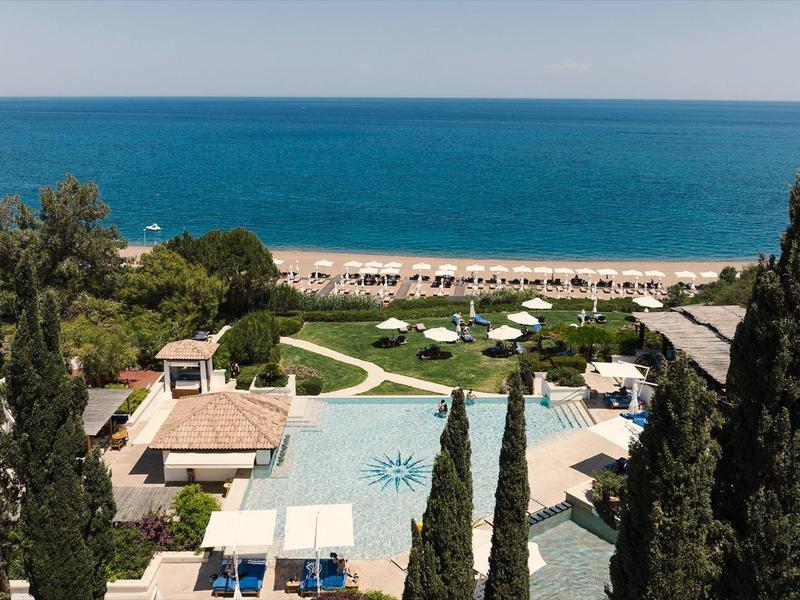 Blick auf einen Hotelpool mit Liegestühlen, Zypressen und dahinter das blaue Meer unter klarem Himmel.