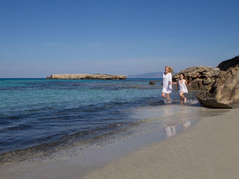 Zwei Personen am Sandstrand neben Felsen, klarer blauer Himmel und ruhiges Meer mit Insel im Hintergrund.