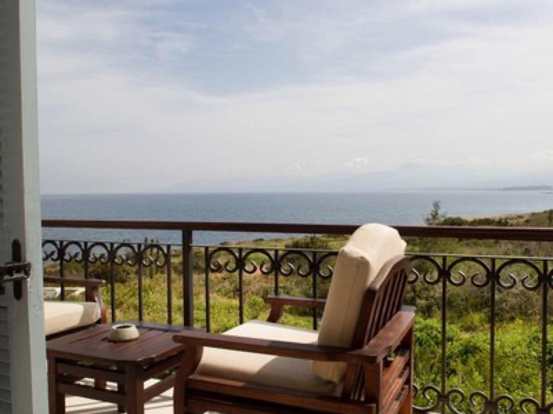 Balkon mit beige gepolstertem Holzstuhl, Holztisch und Blick aufs Meer unter bewölktem Himmel.