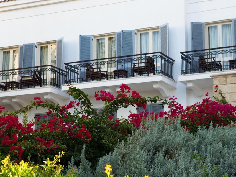 Haus mit weißen Wänden, blauen Fensterläden und Balkonen, davor blühende rote und grüne Pflanzen.