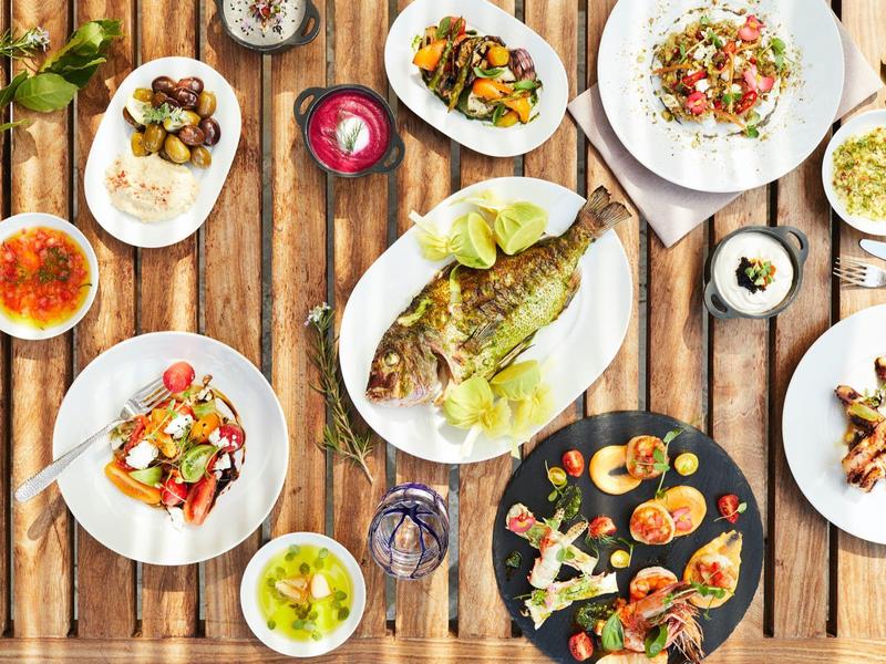 Verschiedene bunte Gerichte mit Fisch, Salaten und Beilagen auf rustikalem Holztisch angerichtet.