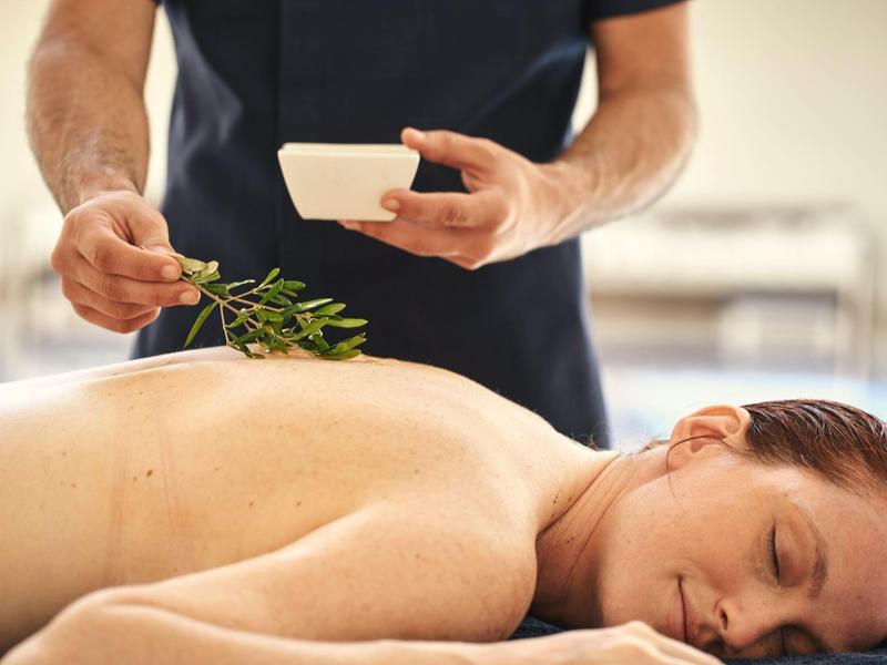 Frau liegt entspannt auf Massagetisch, Mann hält ätherisches Öl und frische Pflanze bereit.