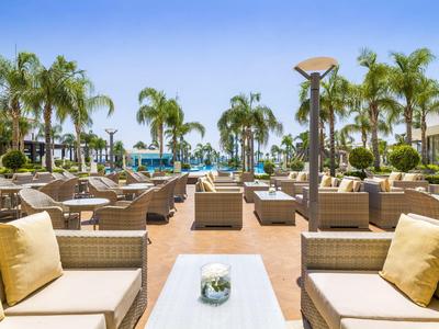 Außenbereich mit beigen Sitzmöbeln und Palmen vor blauem Himmel und Pools.