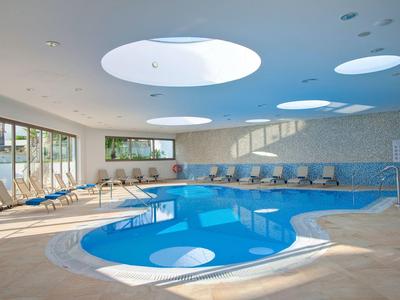 Innenraum mit ovalem Hallenbad, großen runden Oberlichtern und bequemen Liegestühlen am Rand.