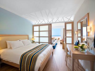Helles Hotelzimmer mit großem Bett, Holzfußboden, Schreibtisch, TV und Blick auf das Meer.