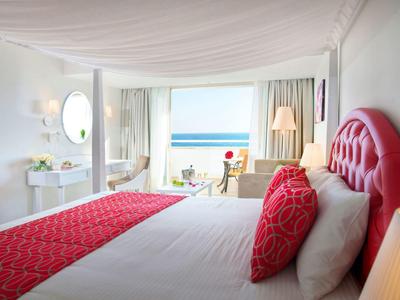 Helles Hotelzimmer mit großem Bett, roten Akzenten und Meerblick durch bodentiefe Fenster.