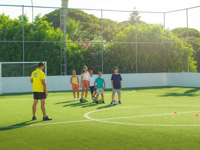 Pięć osób, czworo dzieci i dorosły, stoi na nasłonecznionym boisku sportowym z zieloną sztuczną nawierzchnią.