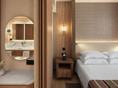 Nowoczesny pokój hotelowy z łóżkiem podwójnym, drewnianymi meblami i podświetlanym lustrem w łazience.