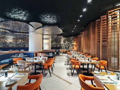 Nowoczesna restauracja hotelowa z okrągłymi i prostokątnymi stołami, pomarańczowymi krzesłami i stylowym oświetleniem.