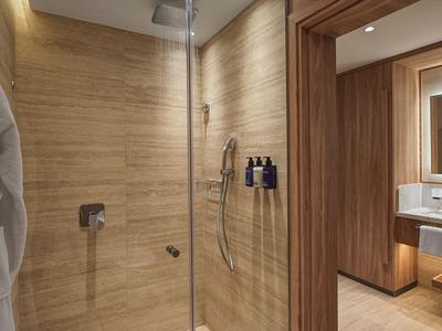 Nowoczesna łazienka hotelowa z prysznicem typu walk-in, drewnopodobne płytki i białe ręczniki.