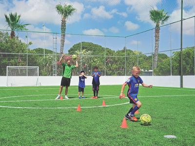 Kinder spielen Fußball auf einem eingezäunten Fußballfeld mit einem Trainer.