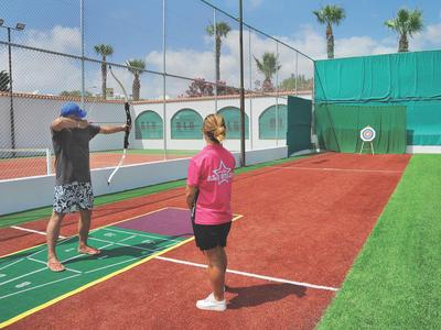 Zwei Personen spielen Bogenschießen auf einem Sportplatz mit Kunstrasen und Tennisplatz im Hintergrund.