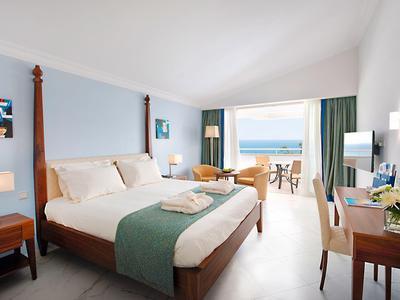 Helles Schlafzimmer mit Doppelbett, Holzmöbeln und Blick auf das Meer durch große Fenster.