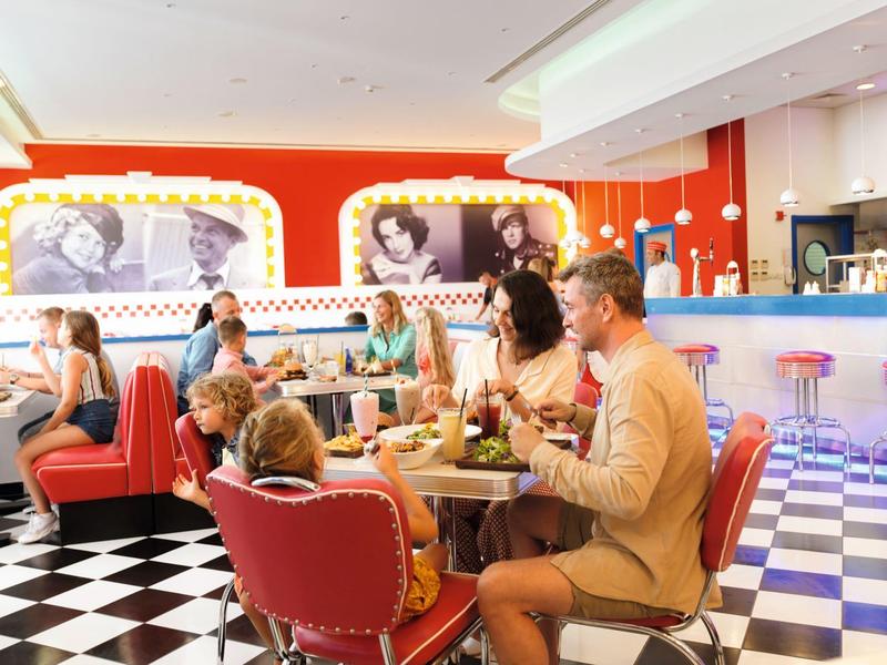 Bunte Retro-Diner mit rot-weißen Sitzecken und schwarz-weiß kariertem Boden. Menschen essen und sprechen.