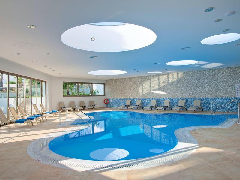 Innenraum mit ovalem Hallenbad, großen runden Oberlichtern und bequemen Liegestühlen am Rand.