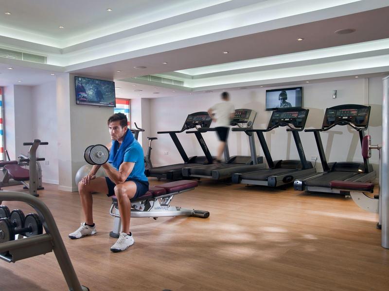 Mann in blauem Shirt sitzt auf Hantelbank in hellem Fitnessraum mit Laufbändern und TV-Bildschirmen.