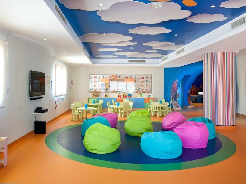 Bunter Kinderraum mit Sitzsäcken, Holzmöbeln, Wandbild und Decke mit Wolken- und Sonnenmotiven.