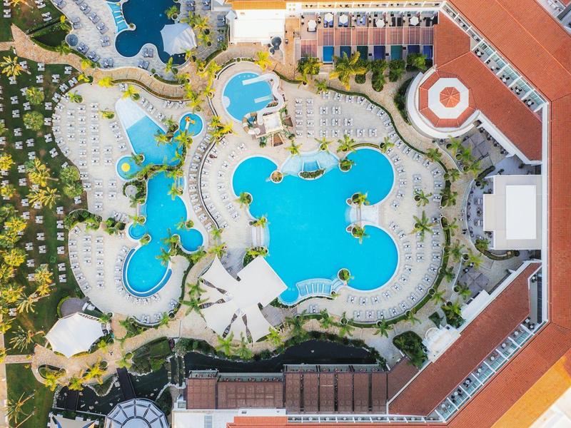 Großer Hotelpool mit mehreren blauen Becken, Liegestühlen und umliegenden roten Dächern und Pflanzen.