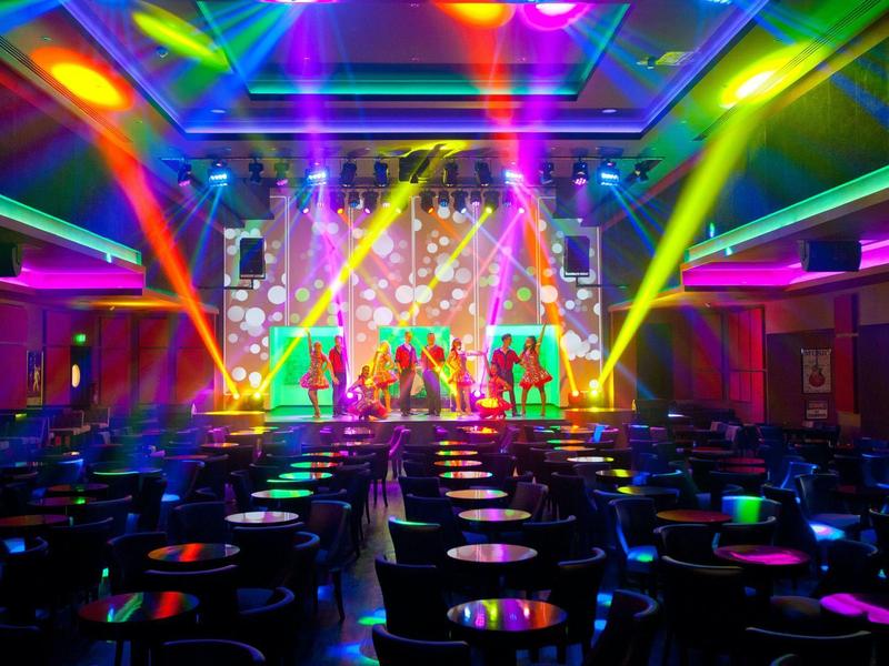 Bunte Bühne mit tanzenden Personen und leuchtenden Scheinwerfern in einem großen Saal.