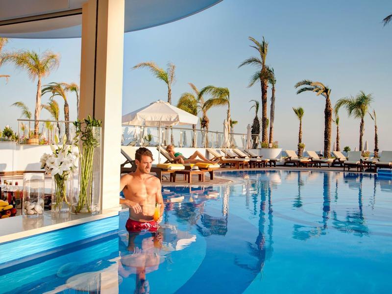 Mann im blauen Pool mit Cocktail am Barbereich, Palmen und Liegen unter Sonnenschirmen bei blauem Himmel.