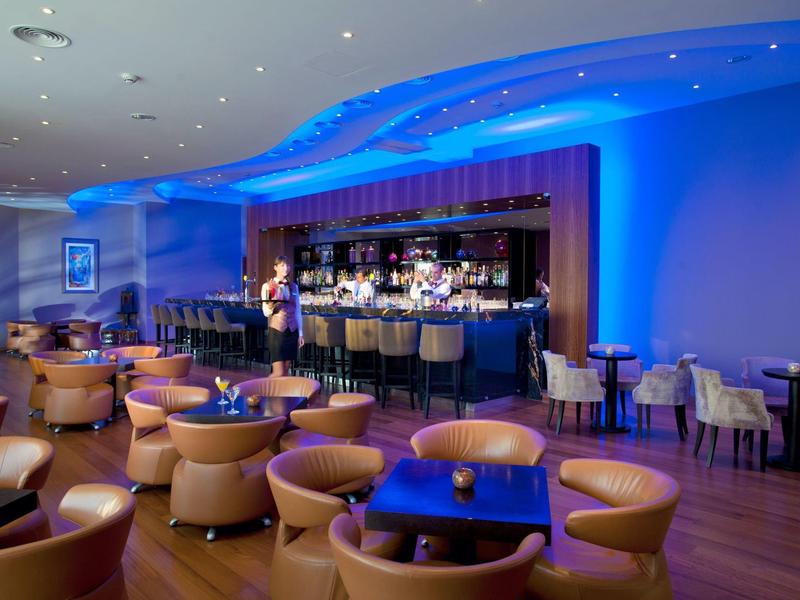 Moderne Bar mit runden Tischen, bequemen Sesseln und blaublauer Akzentbeleuchtung an der Decke.