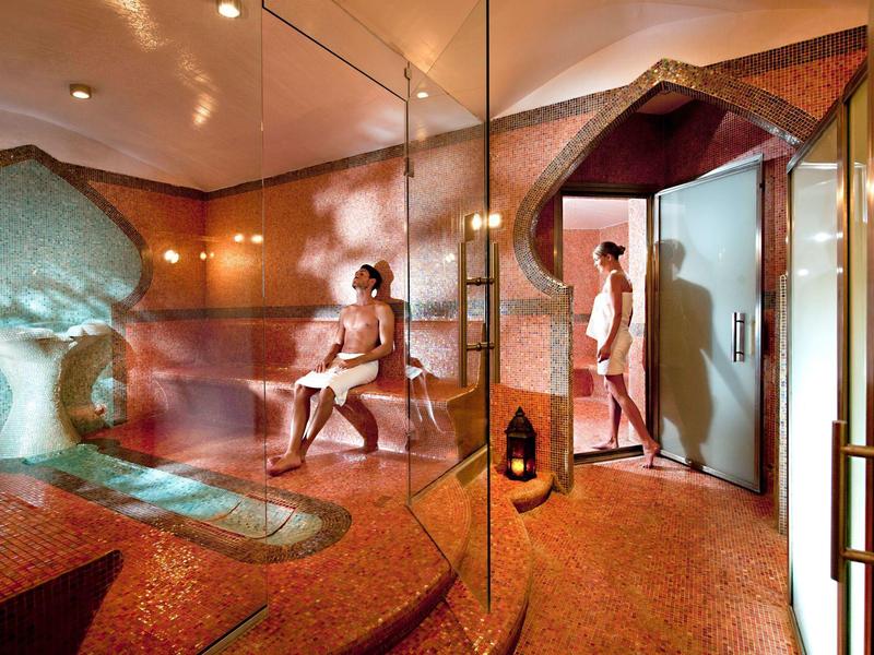 Helle Sauna mit Glastüren, Holzbank, orangem Boden und zwei nackt sitzenden Personen.