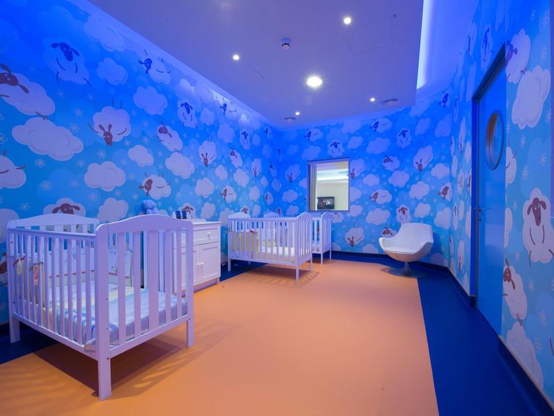 Kindergartenraum mit blauen Wänden, weißen Wolken, 3 Gitterbetten und hellorangefarbenem Boden.