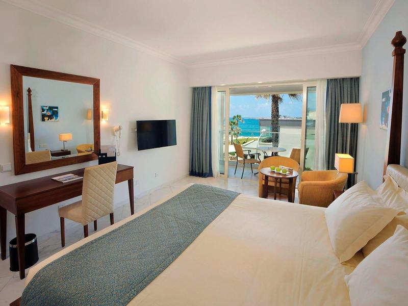 Helles Hotelzimmer mit großem Bett, Schreibtisch, Meerblick und Balkon mit Sitzbereich.