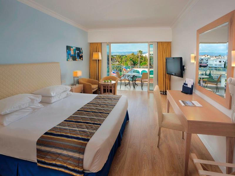 Hell eingerichtetes Hotelzimmer mit Doppelbett, Schreibtisch, Fernseher und Balkon mit Blick auf Pool und Meer.