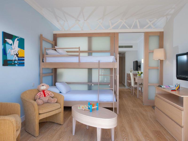 Kinderzimmer mit Etagenbett, zwei Sesseln, kleinem Tisch, Kommode und Fernseher, helle Holzmöbel.